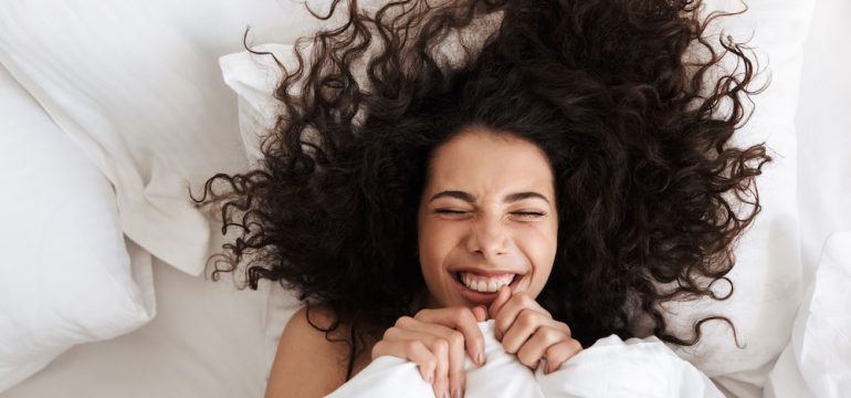 Portrait from above of cheerful woman with dark curly hair and a smiling face lying in bed under white bed sheets pulled up to her chin.