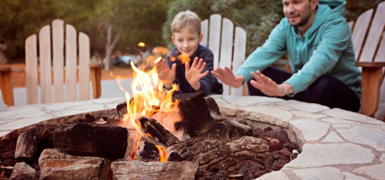 View of firepit and happy smiling family of two, father and son, warming their hands by the fire and enjoying time together in the background.