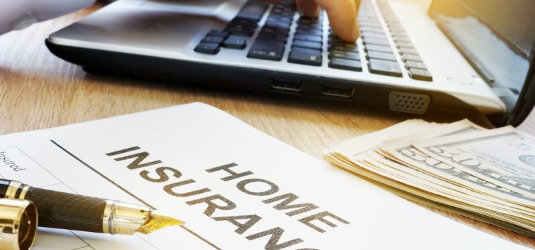 Homeowners insurance form on a table with an agent's hands typing on a laptop computer. A stack of money is also on the desk to represent money saved on homeowners insurance.