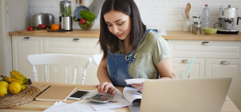 Indoor shot of attractive young female doing calculations, managing monthly bills, sitting at kitchen table and smiling.