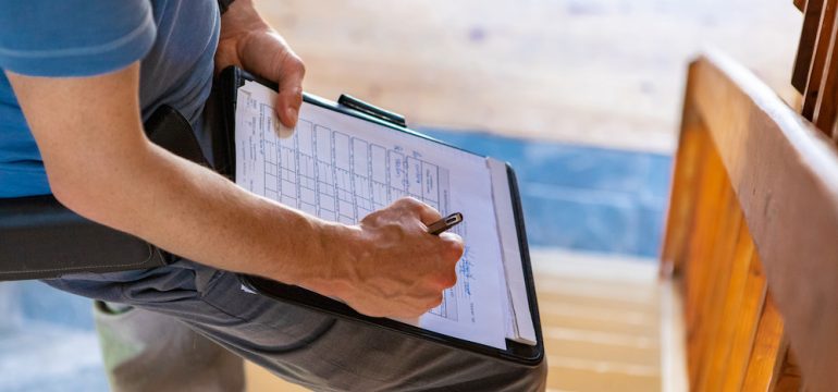 A close up and high angle view of a professional male wearing blue t-shirt, writing out forms during a FHA home inspection, standing on stairs with copy-space.