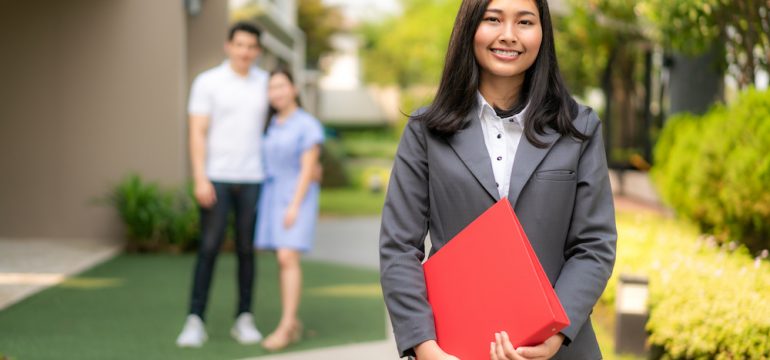 Female real estate agent in a suit holding red file and smiling with a young couple buying a home in front of a house.