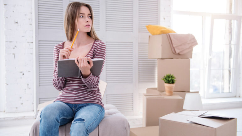 Neat and organized young woman sitting on an ottoman and reviewing a moving checklist.