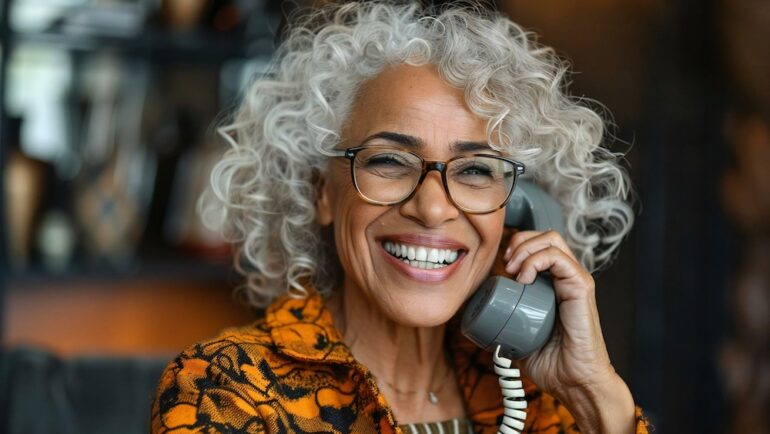 Senior woman smiling and laughing while having a conversation on a landline phone, wearing eyeglasses and looking beautiful. Example of a typical user of landlines in their home.