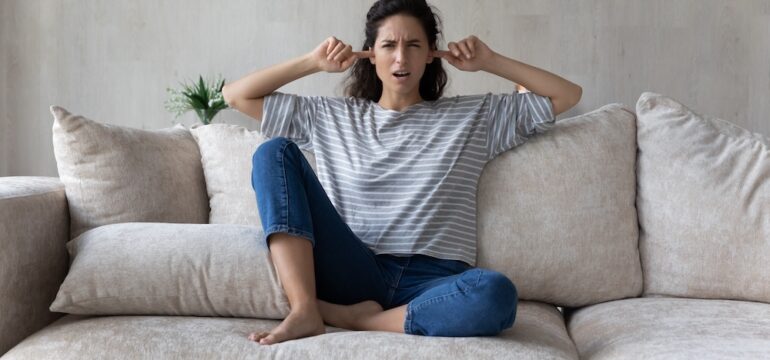 Unhappy woman plugging ears with fingers, sitting on couch at home. Annoying neighbor behaviors concept. Irritated young female ignoring avoiding loud sound of neighbors.